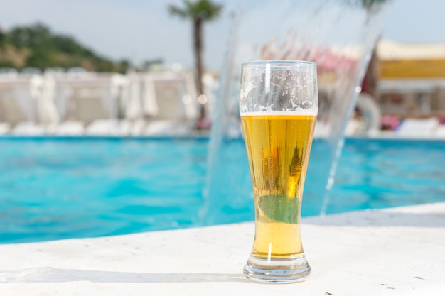 Verre de bière glacée debout sur les carreaux au bord d'une piscine bleu turquoise dans un complexe tropical conceptuel de vacances d'été