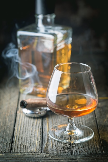 Verre et bouteille de cognac ou de cognac et cigare fumant sur la table en bois.