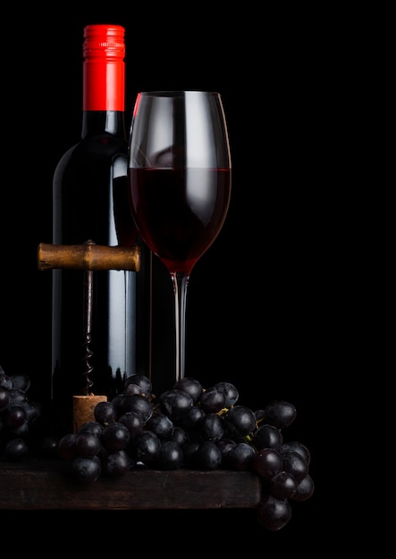 Verre et bouteille de vin rouge avec des raisins noirs et ouvre-tire-bouchon vintage et bouchon sur planche de bois sur fond noir