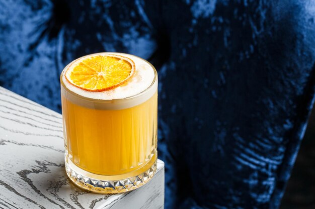 Un verre d'un cocktail jaune avec un gros glaçon en mousse blanche décoré de copeaux d'orange sur l'arrière-plan flou bleu