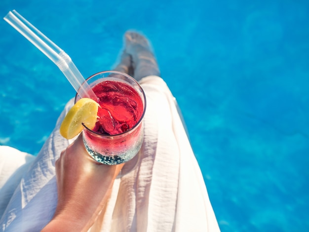 verre avec un cocktail tenu par une personne à côté d'une piscine