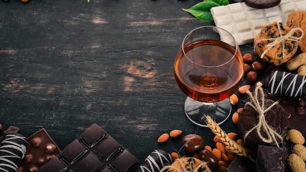 Un verre de cognac brandy whisky et un ensemble de chocolat avec des biscuits et des bonbons sur un fond de bois noir Copiez l'espace pour le texte