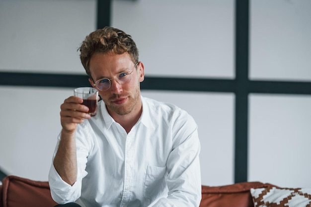 Avec un verre dans les mains Jeune homme caucasien en chemise blanche élégante à l'intérieur à la maison