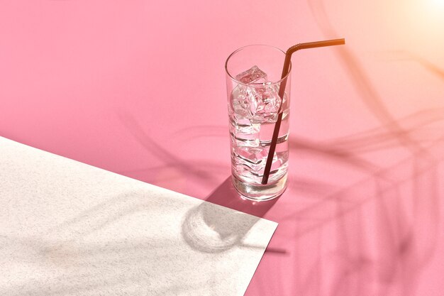 Photo verre d'eau minérale avec de la glace sur fond rose et blanc avec l'ombre d'une fusée solaire en feuille de palmier