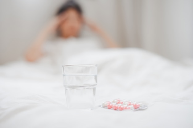 Un verre d'eau et une pilule sur le lit avec une femme malade floue sur fond.