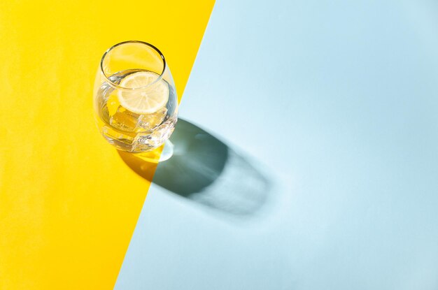 Un verre d'eau avec une tranche de citron et des cubes de glace sur un fond jaune et bleu