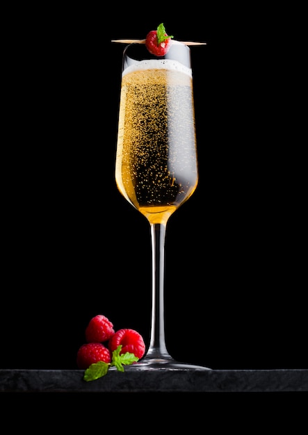 Verre élégant de champagne jaune avec framboise et baies fraîches avec feuille de menthe sur bâton sur panneau de marbre noir sur fond noir.