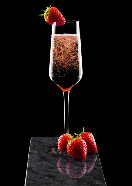 Verre élégant de champagne rose rose avec des fraises sur le dessus et des baies fraîches sur un tableau en marbre noir sur fond noir.