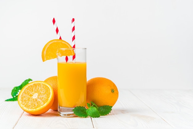 Un verre de jus d'orange fraîchement pressé avec une tranche d'orange et des tubes sur un bureau léger avec des oranges et de la menthe.