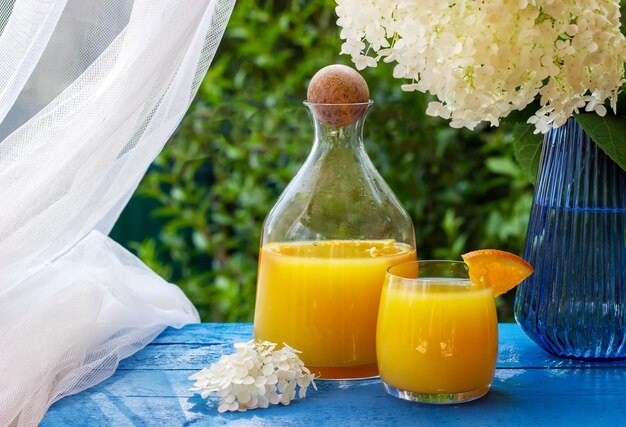 Verre de jus d'orange frais garni d'une tranche d'orange mûre sur la table en bois bleue avec des fleurs en arrière-plan