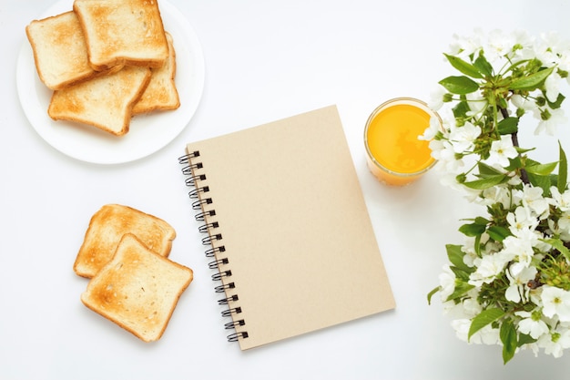Verre de jus d'orange, toasts sur une plaque blanche, bloc-notes et une branche de fleurs sur fond blanc. Concept d'un petit-déjeuner de printemps sain