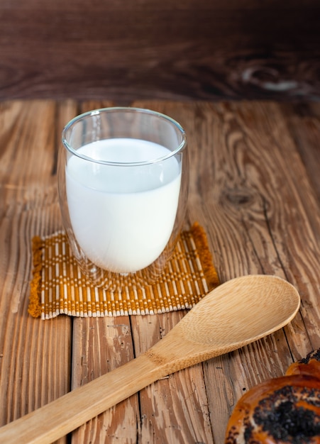 Photo un verre de lait froid avec une cuillère et un petit pain au pavot sur une table en bois