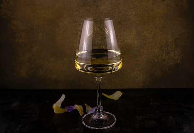 Un verre de vin blanc sur une longue jambe fine avec des pétales de fleurs exotiques sur fond sombre