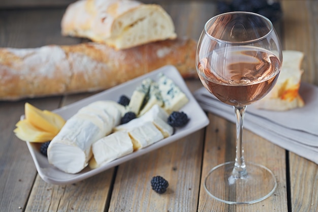 Un verre de vin rosé servi avec assiette de fromages, mûres et baguette. Assortiment de fromages aux fruits rouges sur fond de bois.