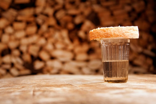 Un verre de vodka et un morceau de pain sur une table en bois et du bois de chauffage derrière