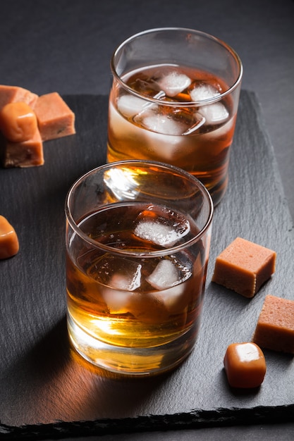 Verre de whisky avec de la glace et des bonbons au caramel sur une ardoise noire sur fond noir