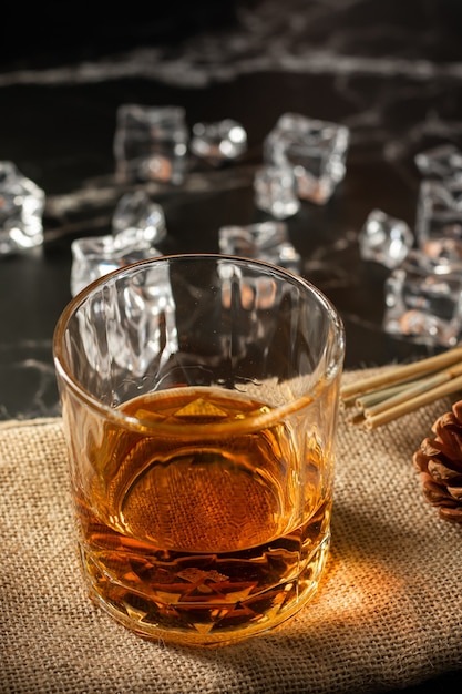 Photo un verre de whisky avec de la glace sur un fond noir en marbre. un verre de whisky écossais et de glace se trouve au sommet d'un baril de whisky rustique, photo verticale.