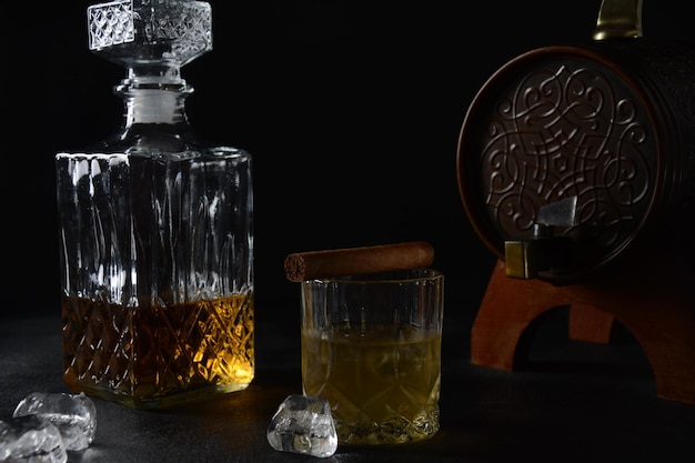 Verre de whisky avec glaçons et carafe carrée, et cigare sur fond noir.