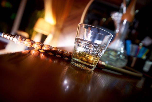 Verre de whisky incliné sur le bar avec un narguilé sur un arrière-plan flou.