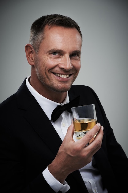 Verre de whisky smoking et portrait d'homme avec de l'alcool dans un costume se sentant chic avec une boisson de luxe Fond gris isolé et studio avec un agent modèle ou un acteur sourire avec une boisson riche