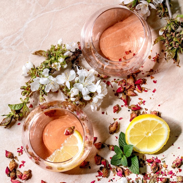 Verres à cocktail de champagne rose rose, de cidre ou de limonade avec des boutons de rose secs, du citron et de la menthe. Branches de cerisier en fleurs au-dessus. Fond de marbre rose. Mise à plat. Image carrée