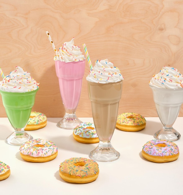 Des verres de délicieux milkshakes crémeux avec des éclaboussures colorées et des pailles beaucoup de bonbons brillants des beignets sur une table blanche
