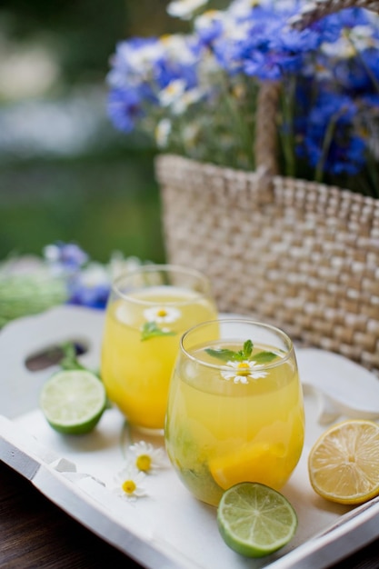 Verres à limonade avec des citrons à la menthe et à l'orange sur un plateau en bois blanc près d'un sac de paille avec des fleurs sauvages Concept de boissons d'été