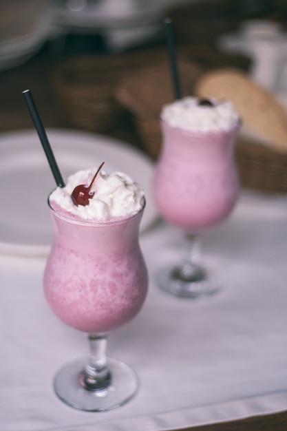 Verres partiellement flous avec milkshake aux baies avec pailles, crème fouettée et cerises cocktail, sur table en bois avec nappe blanche. Verticale