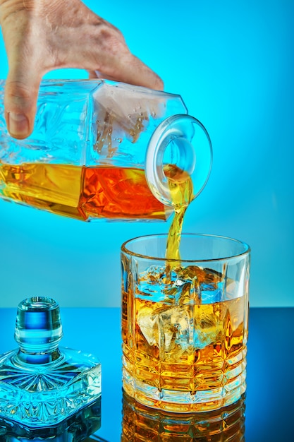 Verser la carafe en cristal carré avec du scotch whisky ou du brandy dans un verre rond en cristal sur un fond dégradé bleu avec reflet