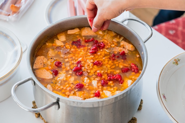 Verser à la main de la framboise dans une soupe de saumon avec des carottes et des légumes dans un pot en acier inoxydable de cuisine traditionnelle