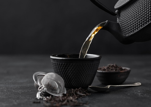 Verser le thé noir de la théière à la tasse avec infuseur à tamis et thé en vrac dans un bol en céramique sur fond noir