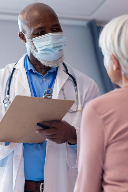 Vertical d'un médecin masculin senior diversifié portant un masque facial parlant avec une patiente âgée, espace de copie. Services hospitaliers, médicaux et de santé.
