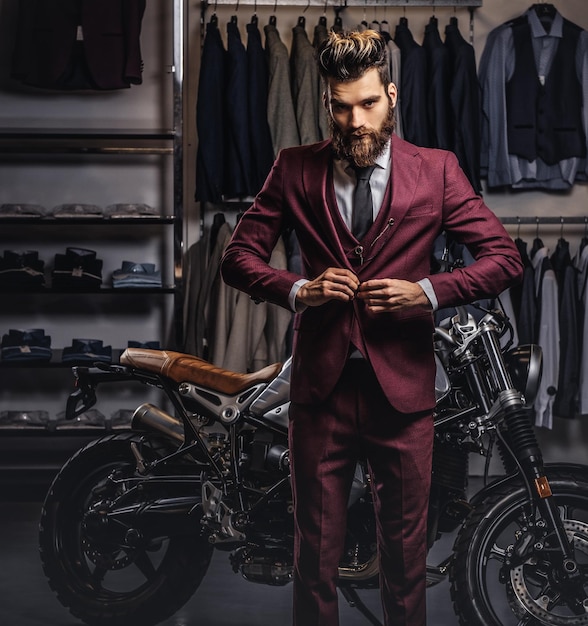 Veste à boutons homme barbu élégamment vêtue près d'une moto de sport rétro debout dans un magasin de vêtements pour hommes.