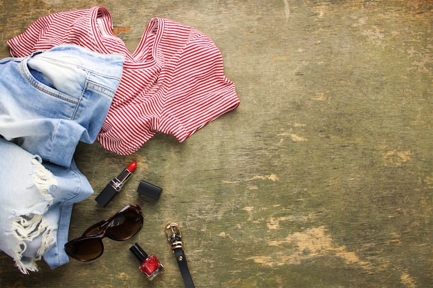 Vêtements et accessoires d'été pour femmes: t-shirt, jeans, rouge à lèvres, vernis à ongles, ceinture, lunettes de soleil. Vue de dessus.