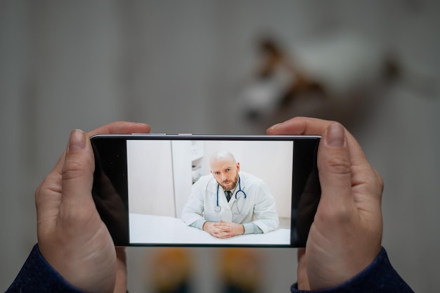 Le vétérinaire conseille le propriétaire du chien en ligne sur son mobile Une femme parle à un médecin lors d'un chat vidéo à propos d'un animal de compagnie Gros plan sur des mains féminines avec un smartphone Un vétérinaire masculin traite un chiot