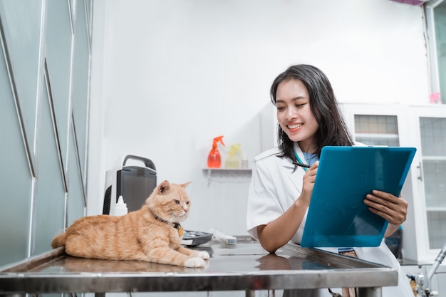 Vétérinaire souriant tout en écrivant sur le presse-papiers quand regarde un chat est assis sur la table à la clinique vétérinaire