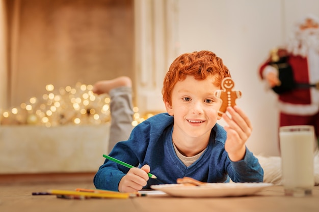 En veux-tu un. Détendu petit garçon souriant tout en tenant un homme en pain d'épice cuit au four et en dessinant sur le sol.