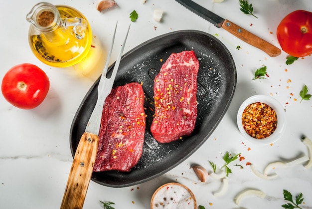 Viande, boeuf. Steaks crus frais dans une poêle. Épices (sel, poivre), légumes frais tomates, carottes, ail, oignons. Sur une table en marbre blanc, avec une fourchette à viande et un couteau. Vue de dessus