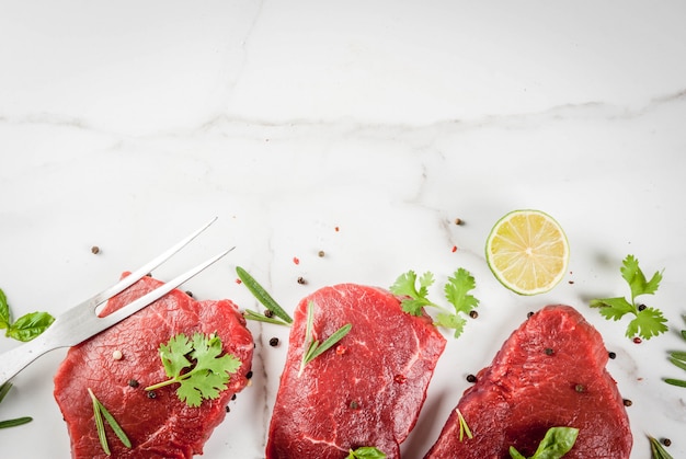 Viande crue fraîche. Filet de boeuf, steaks, sur une table en marbre blanc. Avec de l'huile d'olive, des épices pour la cuisson - basilic, romarin, coriandre, persil, ail, citron, sel, poivre. Copier la vue de dessus de l'espace