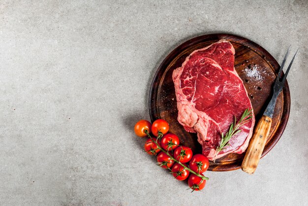 Viande crue fraîche, steak de marbre de boeuf d'agneau sur une planche à découper, avec des ingrédients pour la cuisson. Sur une table en pierre grise, vue de dessus du fond