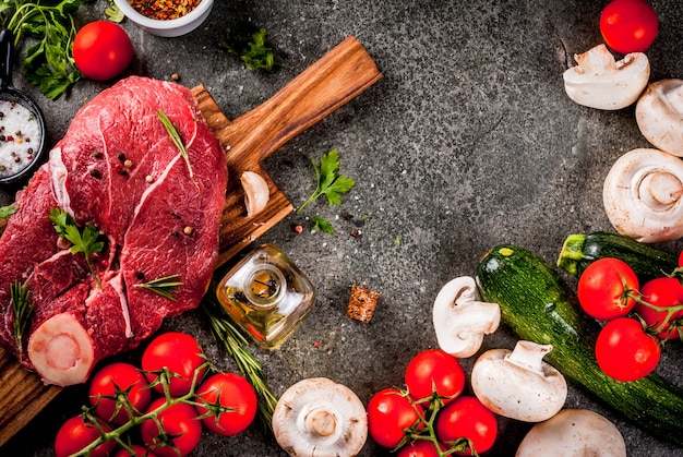 Viande crue avec des ingrédients pour le dîner. Filet de boeuf, filet, sur planche à découper, avec sel, poivre, persil, romarin, huile, ail, tomate, champignon.Table en pierre noire, copie espace vue de dessus