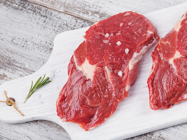 Viande crue. Steak de boeuf frais cru sur une planche à découper blanche avec du romarin et des épices. Fermer