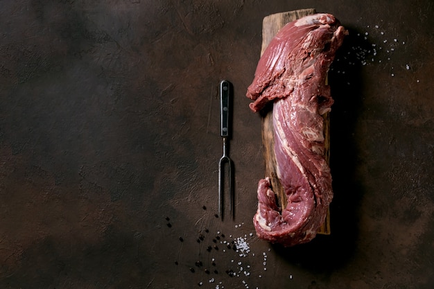 Viande fraîche entière de filet de boeuf cru sur planche de bois avec fourchette à viande en métal, sel et poivre sur une surface de texture brun foncé. Concept de fond de cuisson des aliments. Vue de dessus, mise à plat, espace de copie