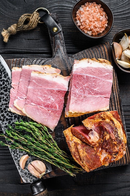 Viande de longe de porc en tranches fumées sur une planche à découper en bois. Fond en bois noir. Vue de dessus.