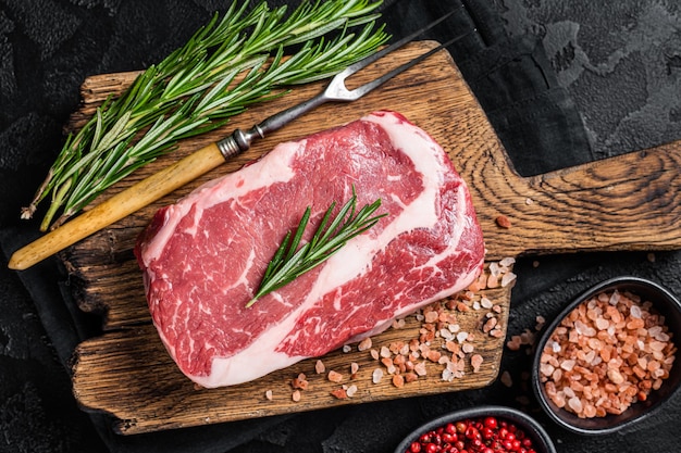 Viande marbrée de boeuf Ribeye Steak cru sur planche de bois avec romarin Fond noir Vue de dessus