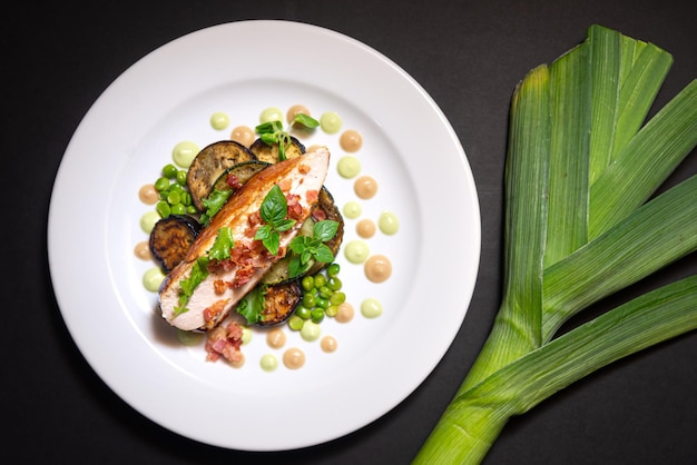 La viande de poitrine de dinde avec des morceaux de bacon frits est assise sur des aubergines cuites au four et des pois verts à côté des jeunes et petites feuilles de basilic et de la purée de chou-fleur et de la purée de pois Sur une assiette blanche et un fond noir