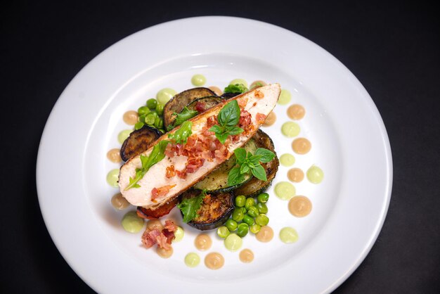 La viande de poitrine de dinde avec des morceaux de bacon frits est assise sur des aubergines cuites au four et des pois verts à côté des jeunes et petites feuilles de basilic et de la purée de chou-fleur et de la purée de pois Sur une assiette blanche et un fond noir