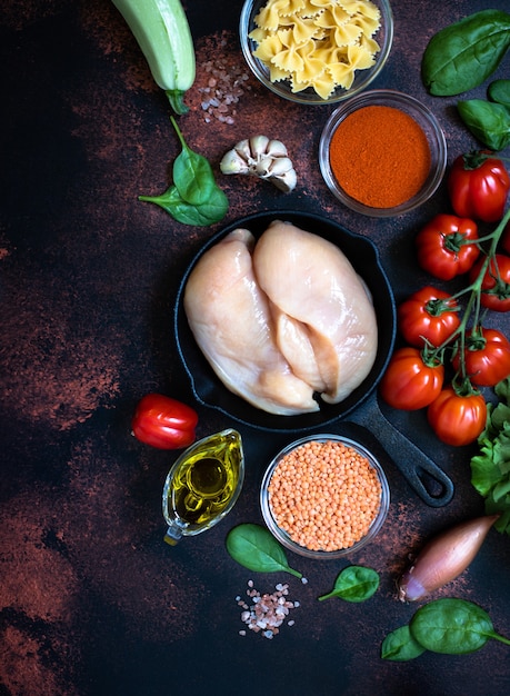 Viande de poulet crue fraîche, filet de poulet dans une poêle entourée d'ingrédients alimentaires sains sur un fond rustique sombre. Vue de dessus, espace copie