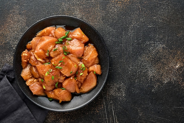 Viande de poulet crue marinée à la sauce soja teriyaki, oignons et poivre dans une assiette noire sur une surface de béton sombre