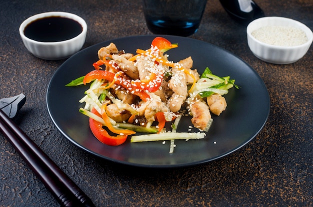 Viande de poulet avec légumes wok, sauces soja et sésame dans une assiette noire avec des baguettes chinoises sur fond sombre. Cuisine asiatique traditionnelle. Espace de copie